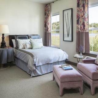 A cozy bedroom featuring a well-made bed, adjacent nightstand with a lamp, and two upholstered armchairs. The room is decorated with floral curtains and art on the wall.
