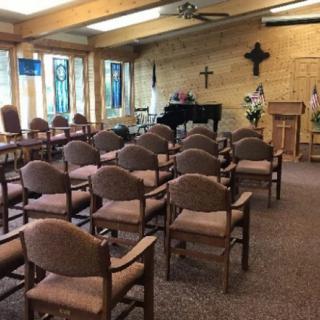A small chapel interior with rows of chairs facing a piano and a wooden pulpit, decorated with crosses and stained glass windows.
