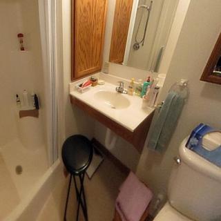 A compact bathroom featuring a sink with toiletries, a mirror, a toilet, a shower area, and a stool.