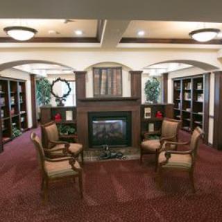 A cozy library reading area with bookshelves and armchairs arranged around a fireplace.