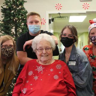 A group of people celebrating Christmas together indoors, with an elderly woman in a red festive sweater at the center.