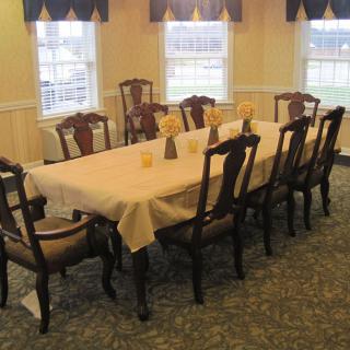 A dining room with a large table and several chairs, illuminated by natural light.