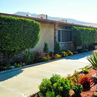 A well-maintained house with neatly trimmed hedges and a colorful garden, set against a backdrop of mountains on a sunny day.