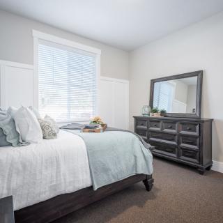 A cozy bedroom with a neatly made bed, a dresser with a mirror, and bright natural lighting.