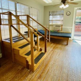 A brightly lit rehabilitation room features a set of wooden stairs and an exercise table.