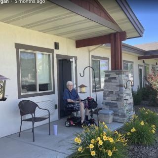 An elderly person sitting on a porch beside flowers on a sunny day.