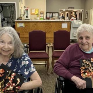Two elderly women in wheelchairs smiling and holding crafts.