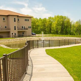 A scenic view of a house by a pond with a pathway and fence, surrounded by a lush lawn and trees on a sunny day.
