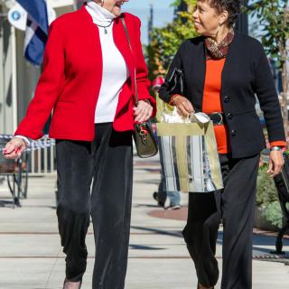 Two women walking and conversing on a city street, one carrying a shopping bag.