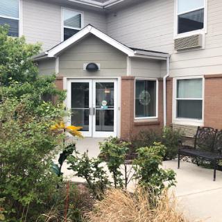 The image shows the exterior entrance of a building with surrounding landscaping and shrubs.