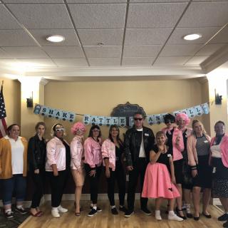 A group of people dressed in 1950s-themed costumes poses under a 'Grease Lightning' banner.