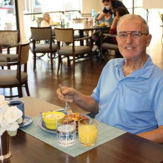 An elderly man enjoying breakfast in a restaurant, smiling while seated at a table with juice and food.