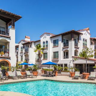 A sunny day at a residential apartment complex featuring a swimming pool surrounded by lounge chairs and umbrellas.