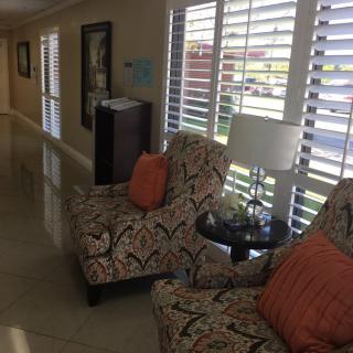 A well-lit lobby area featuring two patterned chairs with orange cushions, a side table with a lamp, and large windows adorned with shutters.