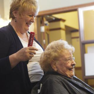 A hairdresser smiling while attending to an elderly woman at a salon.
