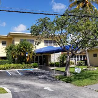 A view of a two-story building with a blue awning, surrounded by greenery and a parking lot.