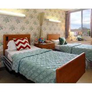 A cozy bedroom with two twin beds, each adorned with blue bedspreads and decorative pillows. The room features a nightstand between the beds and a large window allowing natural light to illuminate the space.