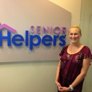 A woman stands next to a company sign that reads 'Senior Helpers' in an office setting.