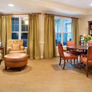 A cozy living and dining area with upholstered chairs, a round dining table, and large windows with green curtains.