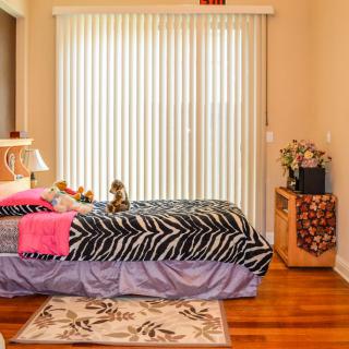A cozy bedroom with zebra print bedding and a floral rug, featuring wooden floors and vertical blinds.