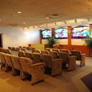 A small auditorium with rows of upholstered chairs facing a wall decorated with stained glass windows and indoor plants.