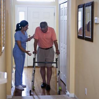 A nurse assists an elderly man using a walker in a home setting.