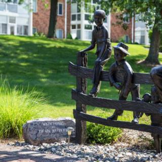 Bronze sculptures of children sitting and playing on a fence in a park setting.