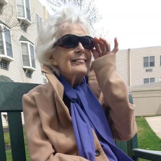 An elderly woman sitting on a bench, wearing sunglasses and a scarf, enjoying a sunny day.