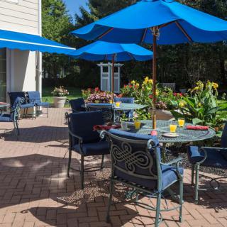 A sunny patio with blue umbrellas and outdoor seating, surrounded by a garden.