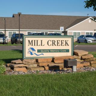 Entrance sign for Mill Creek Nursing Home with building and parked cars in the background.