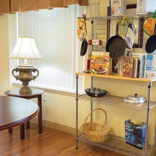 A cozy kitchen corner with a shelf displaying cookbooks and pans, next to a lamp-lit table.