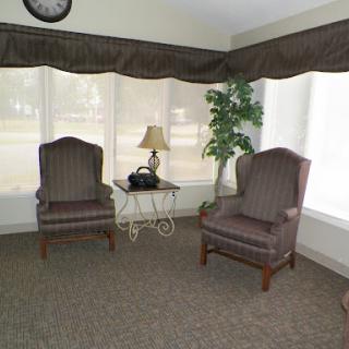 A cozy seating area featuring two chairs, a small table with a lamp, and a plant, illuminated by natural light from large windows.