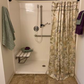 An accessible shower with a folding bench and grab bars, featuring towels and a floral curtain.