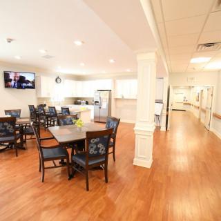 A bright dining area with wooden flooring, featuring tables and chairs
 alongside a modern kitchen.