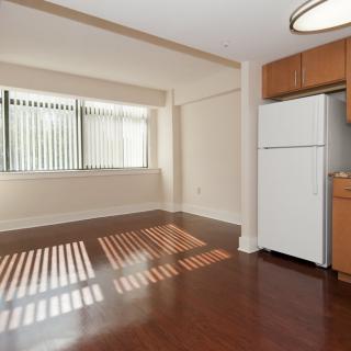 An empty apartment living area with hardwood floors and a kitchen featuring a refrigerator, illuminated by natural light from a large window.