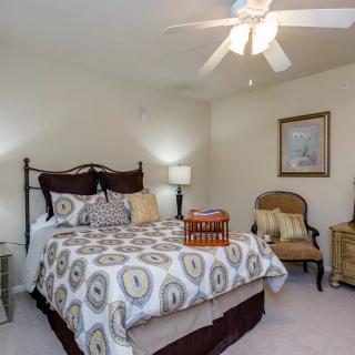 A cozy and well-decorated bedroom featuring a bed with patterned bedding, a side table with a lamp, an armchair, and a ceiling fan.