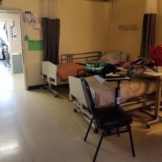 A view of a hospital patient room featuring two beds, a chair, and various medical items.