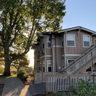 A two-story residential building with outdoor stairs and a large tree in the foreground during sunset.