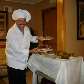 A smiling chef presenting dishes on a table in a kitchen setting.