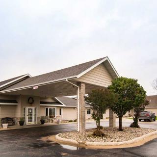 A one-story building with a covered entrance and a neatly landscaped area, including trees and a parking lot, under an overcast sky.