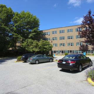 A parking lot with several cars in front of a multi-story building surrounded by trees.