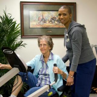An elderly woman exercising on a machine with the assistance of a caregiver in a rehabilitation setting.