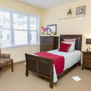 A cozy bedroom with a single bed, featuring red and white bedding, wooden furniture, and natural light from the window.