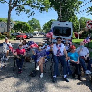 A group of people sitting on chairs along a street, holding American flags during a parade.