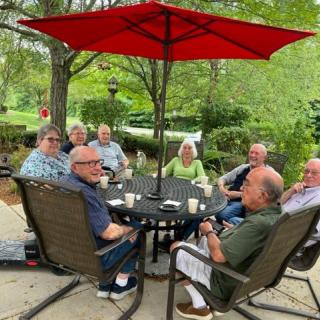 A group of elderly people sitting around a patio table having a conversation under a red umbrella.
