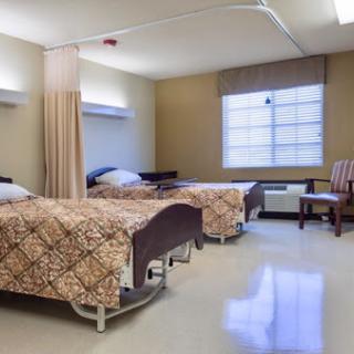 A spacious hospital patient room featuring two beds, a chair, and natural light from a window.
