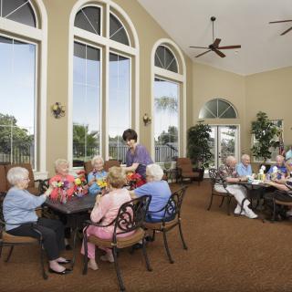 A group of seniors enjoying a social meal in a well-lit dining area with large arched windows.
