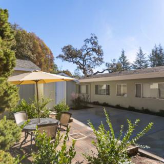 A sunny patio area with outdoor furniture and an umbrella, surrounded by trees and adjacent to a house.