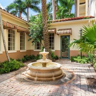 A serene courtyard featuring a central fountain surrounded by tropical plants and palm trees with architectural elements in the background.