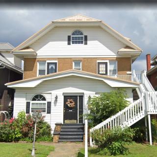 A two-story house with white siding and a decorative archway entrance, surrounded by greenery under a cloudy sky.